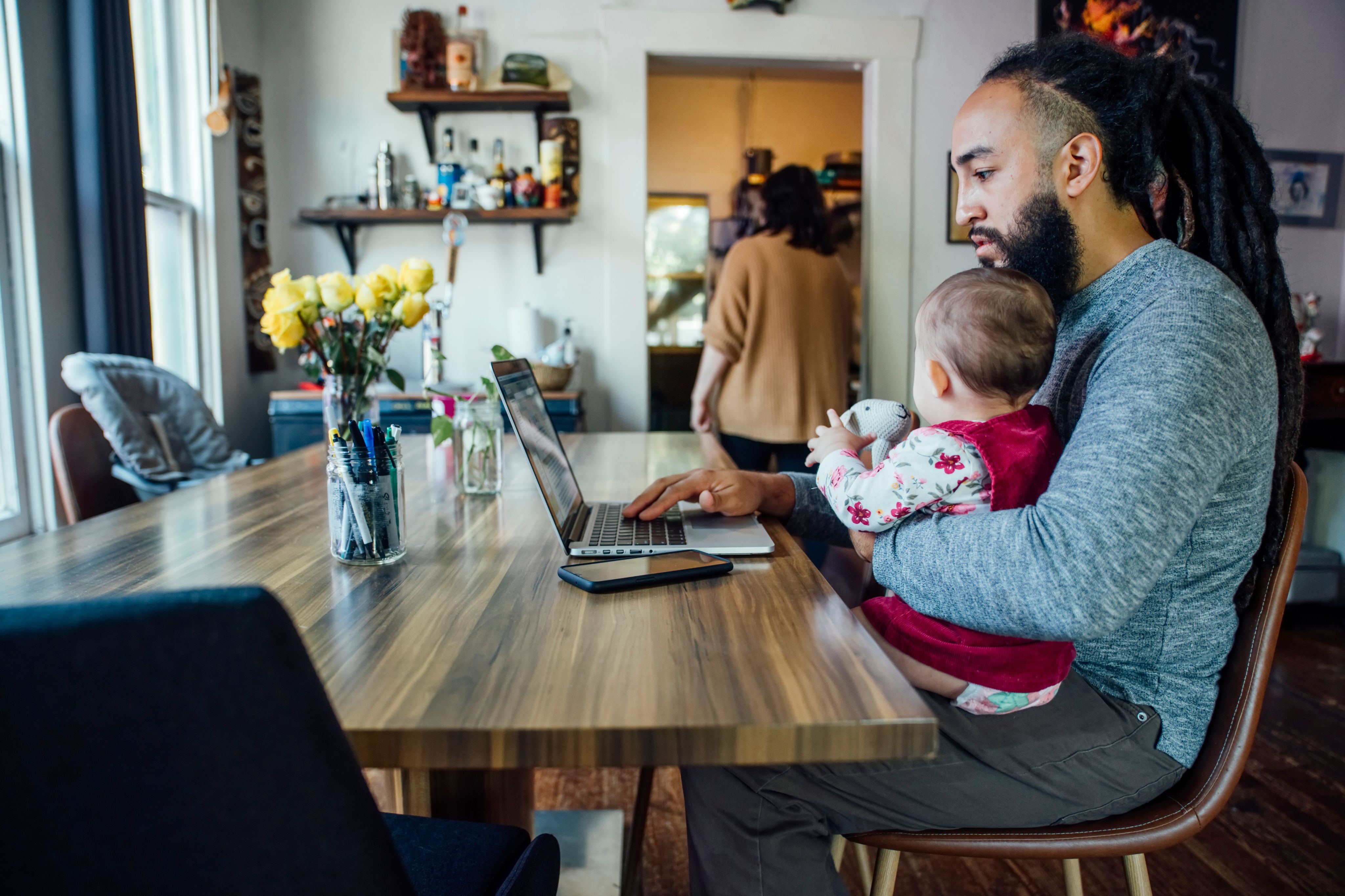 Dad holds child while working from home on laptop at dining room table with partner in background - Copyright: Inti St. Clair / AdobeStock