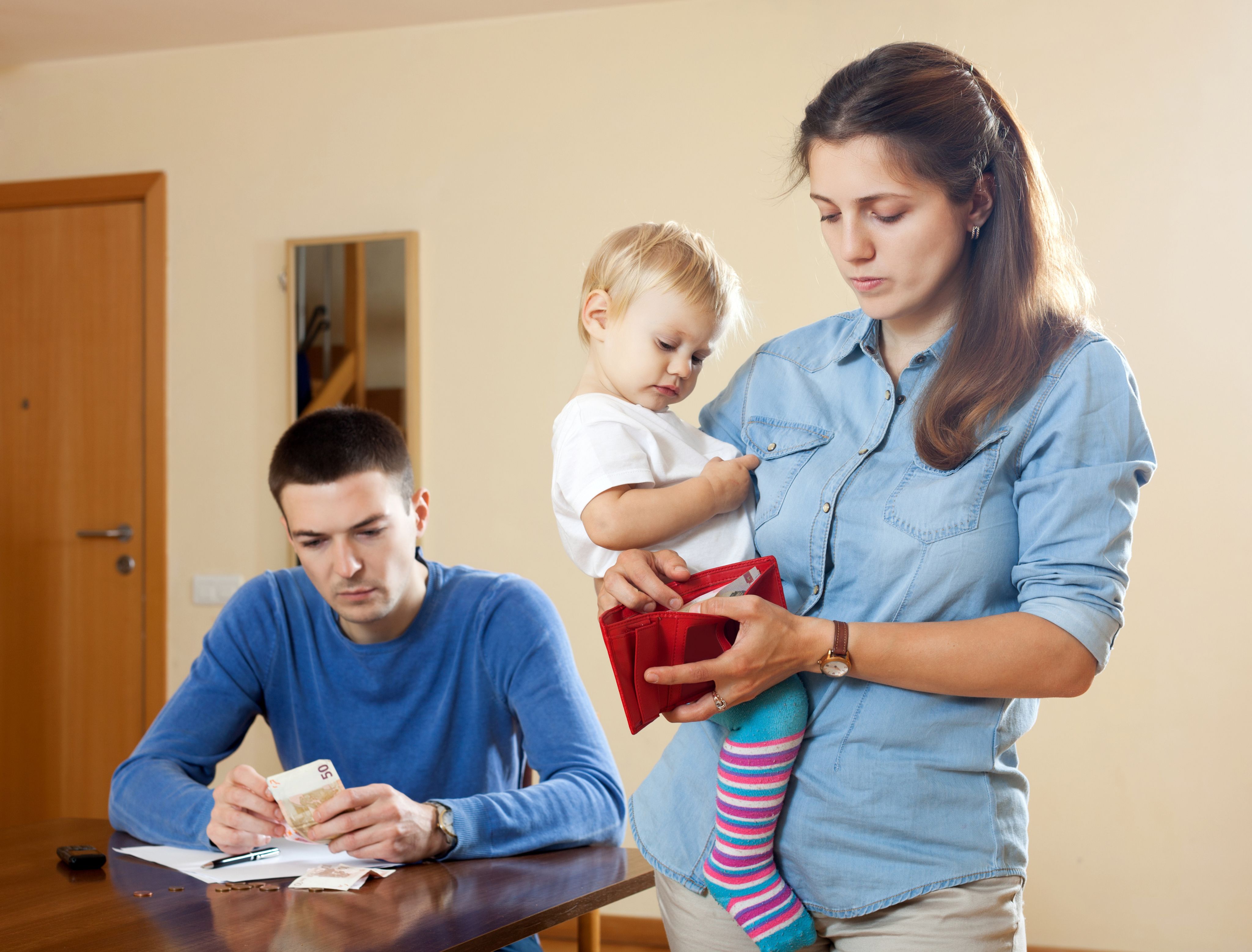 Young mother with toddler in arms looking into her wallet while partner counts out money at the table - Copyright - JackF / Adobe Stock