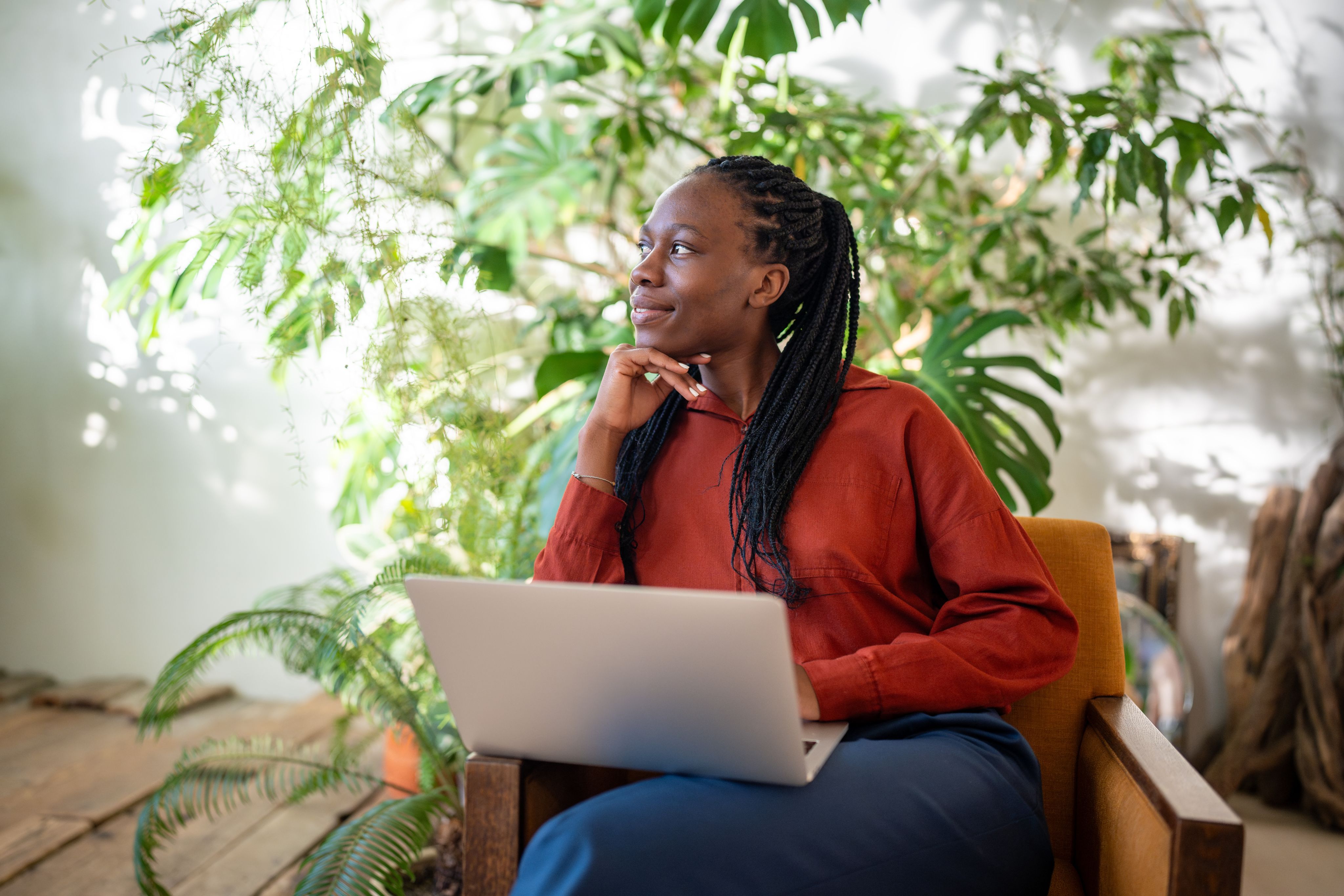 Relaxed black female takes a screen break from computer, looks out the window, surrounded by houseplants. Copyright - DimaBerlin / Adobe Stock
