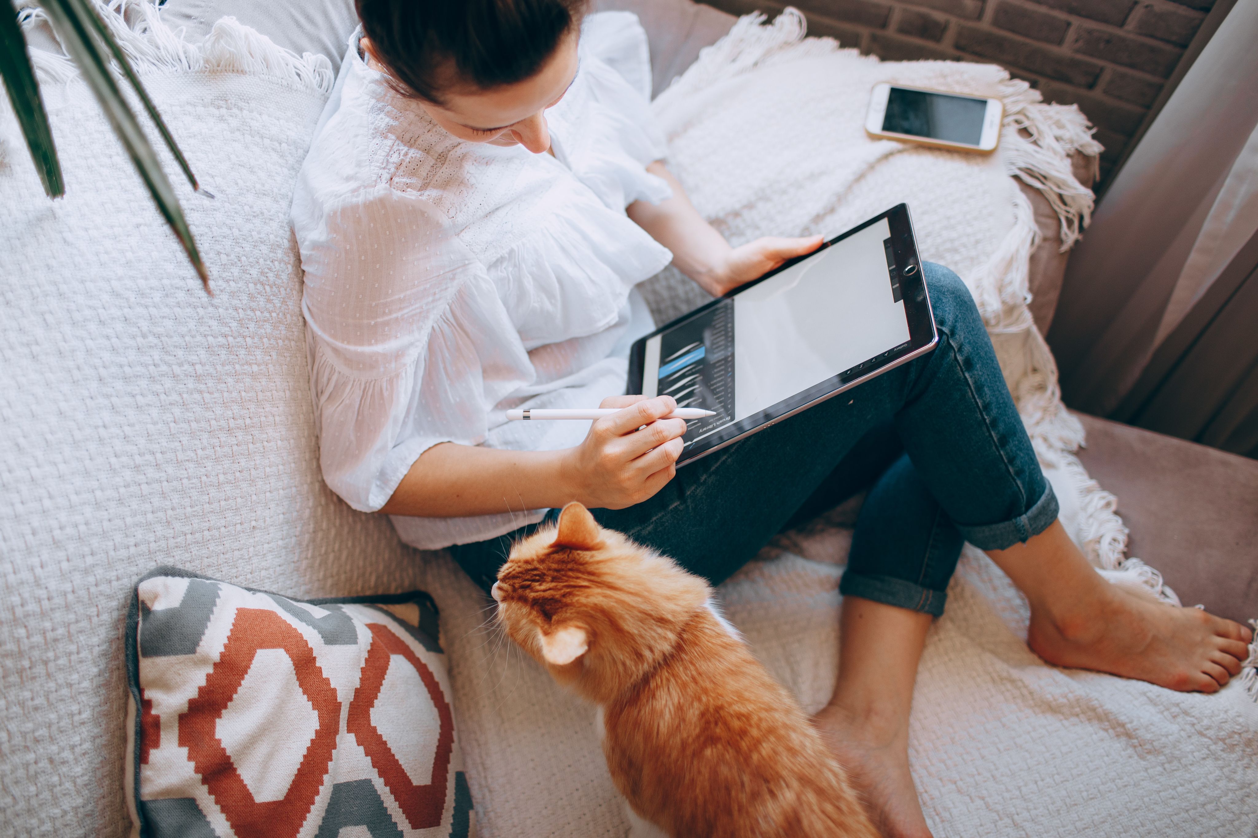woman in white long sleeve shirt and blue denim shorts sitting on bed using tablet computer with kitten